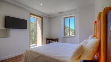 Un dormitorio con cama, televisor montado en la pared y dos ventanas con vistas a zonas verdes y edificios.