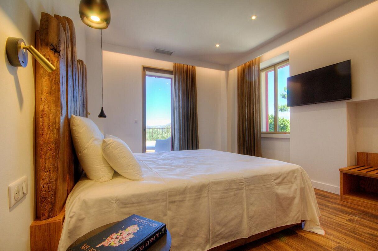 Un dormitorio con una cama grande, cabecera de madera, mesita de noche, televisor montado en la pared y una ventana con cortinas.