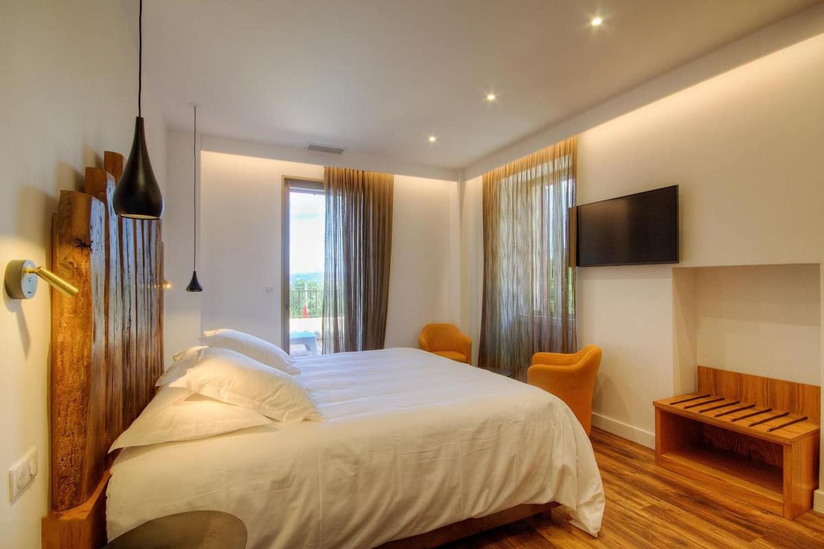 A modern hotel room with a large bed, a wooden bench, a TV mounted on the wall, and a balcony with curtains.