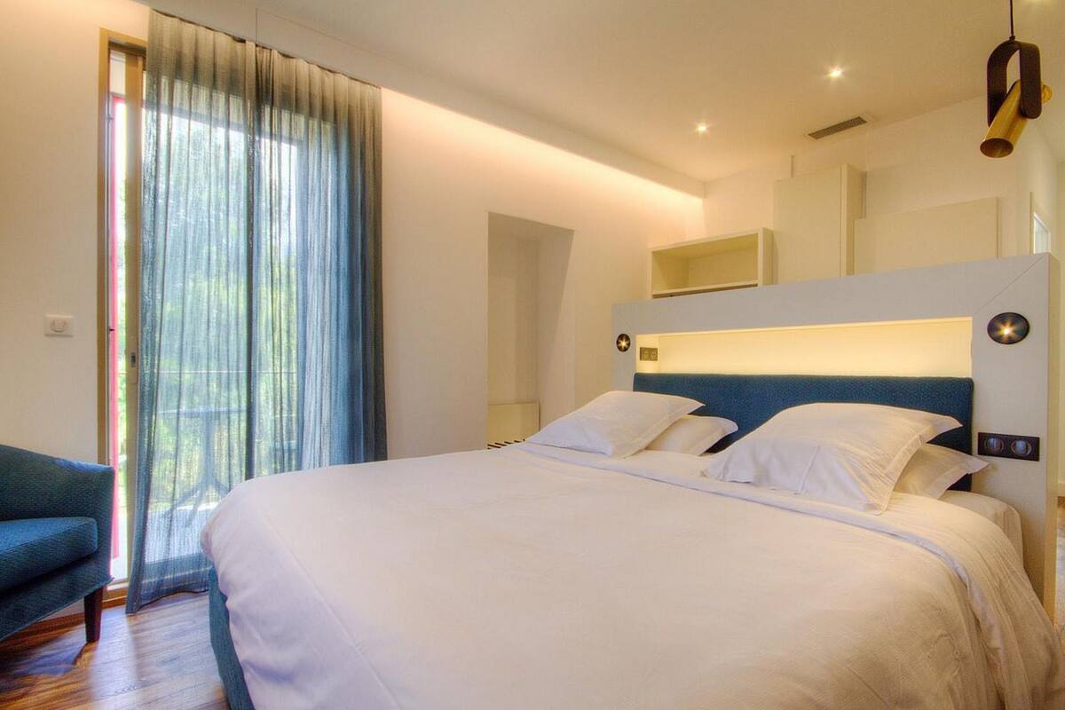 A modern hotel room with a large bed, a sofa, and a view through a window with sheer curtains.