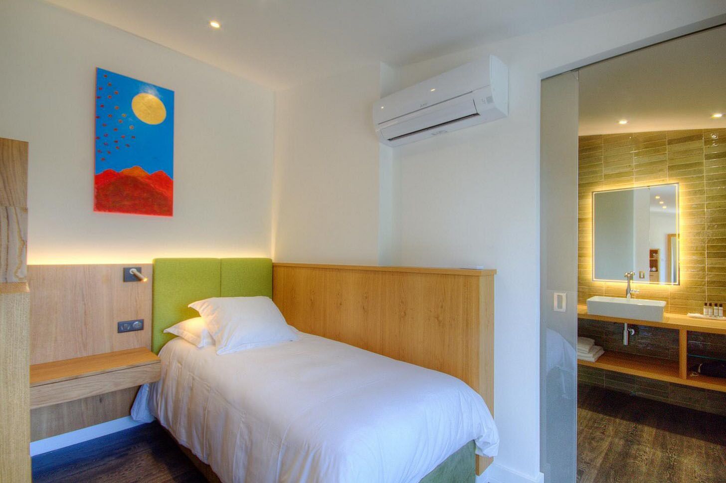 Habitació d'hotel amb un llit, una tauleta de nit, un quadre, un aparell d'aire condicionat i un bany amb un lavabo i mirall.