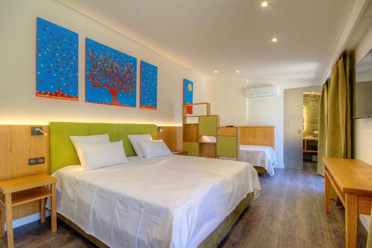 A hotel room with a large bed, a desk, and a chair. There are three paintings on the wall and a window with curtains.