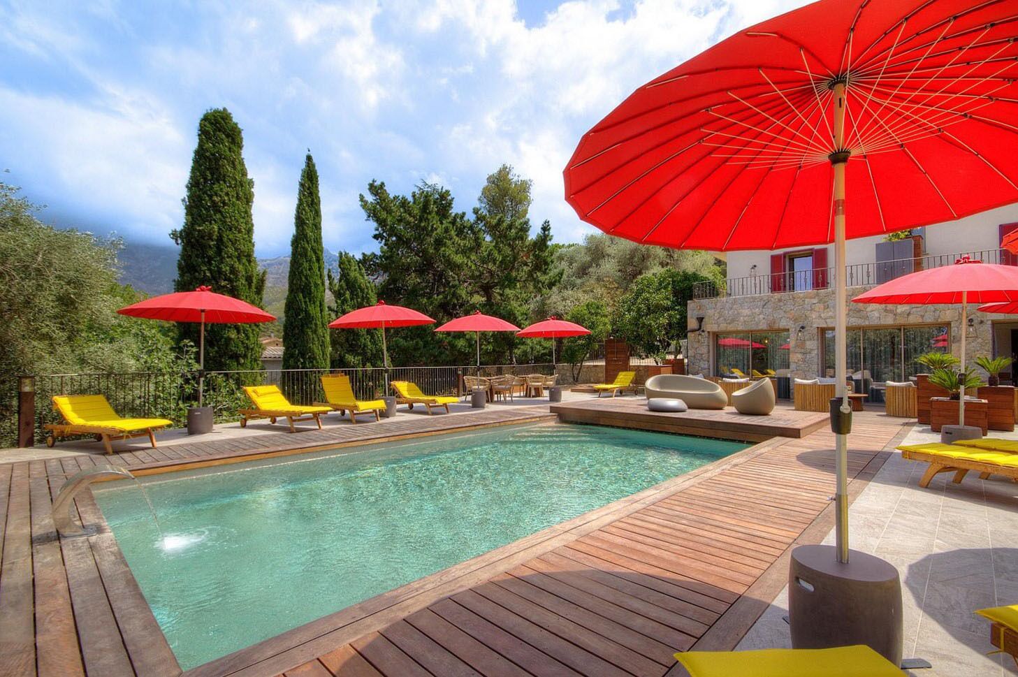 Un espace piscine doté d’une terrasse en bois, de chaises longues jaunes et de parasols rouges.