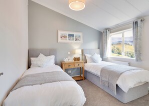 A bedroom with two beds, a nightstand with a lamp, a framed picture on the wall, and a window with a view.