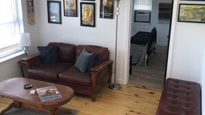 Una sala de estar acogedora con un sofá de cuero marrón, una mesa de centro de madera y un sillón estampado. La habitación tiene piso de madera, un ventilador de techo y varios carteles enmarcados en las paredes.