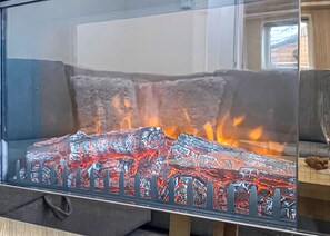 Ein gläserner Kamin mit sichtbaren Flammen, ein graues Sofa und ein Holztisch im Hintergrund.