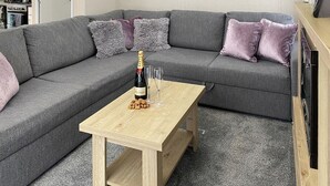 A grey sectional sofa with cushions, a wooden coffee table, and a bottle of champagne. - 2 bedroom accommodation in Snowdonia (Snowdonia)