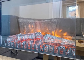 A glass fireplace with visible flames, a grey sofa, and a wooden table in the background.