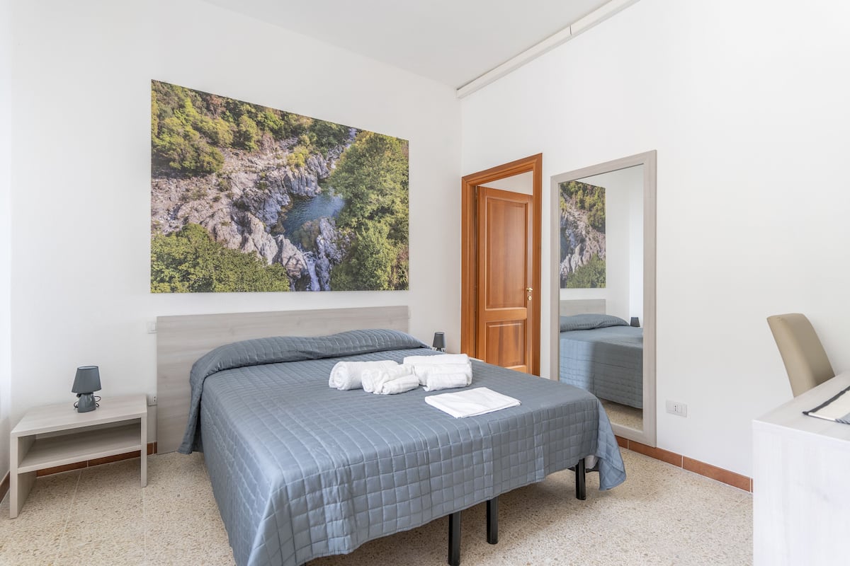 A bedroom with a large bed, a nightstand, a lamp, and a painting of a natural landscape.