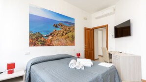 A bedroom with a bed, a desk, a TV, and a painting of a coastal landscape. - Hotel dei Tacchi (Osini)