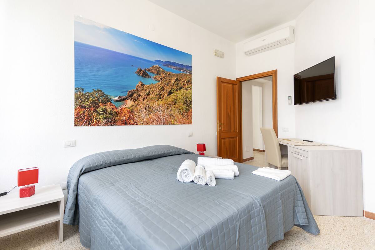 A bedroom with a bed, a desk, a TV, and a painting of a coastal landscape.