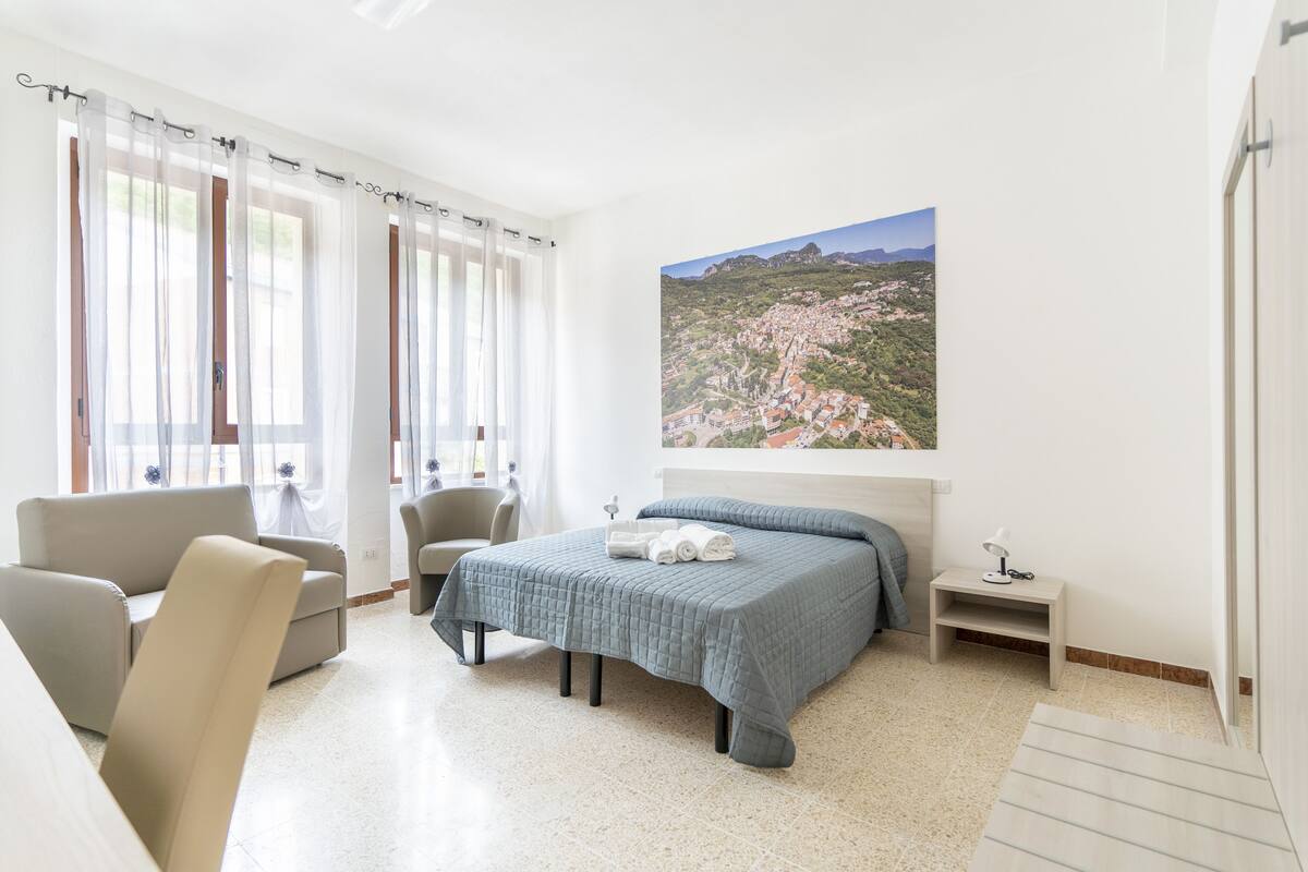 A bedroom with a bed, a chair, a small table, and a painting of a landscape.