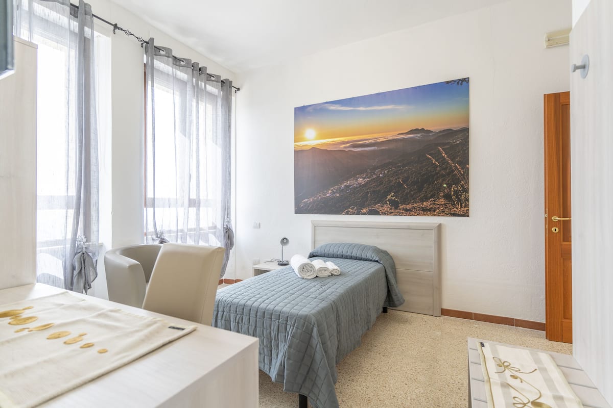 A hotel room with a bed, a desk, a chair, and a large landscape painting on the wall.