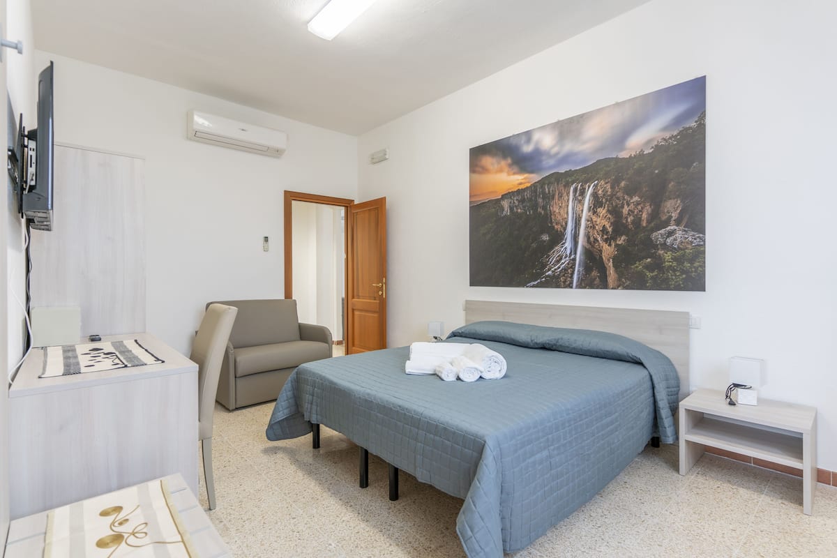 A hotel room with a bed, a desk, a chair, a TV, and a painting of a waterfall.