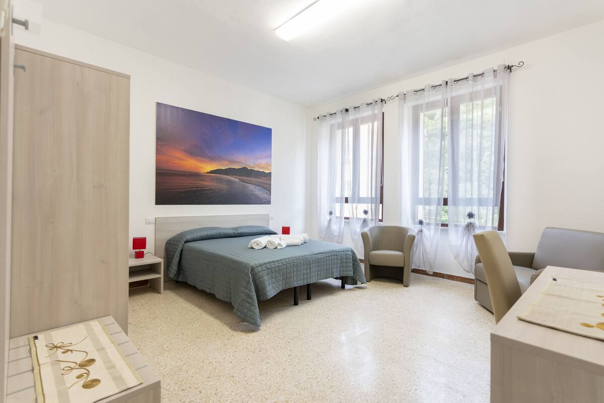 A hotel room with a bed, a chair, a desk, and a painting of a sunset.