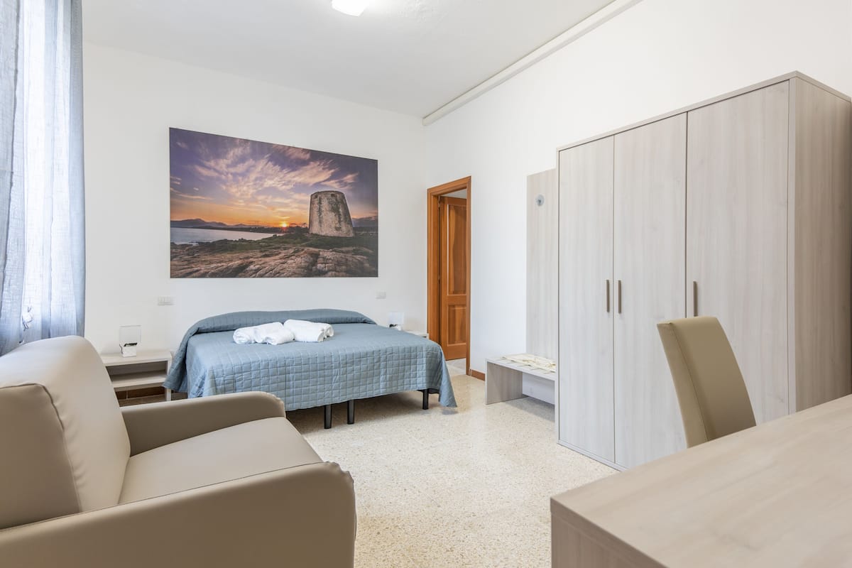 A bedroom with a bed, a desk, a wardrobe, and a painting of a sunset.