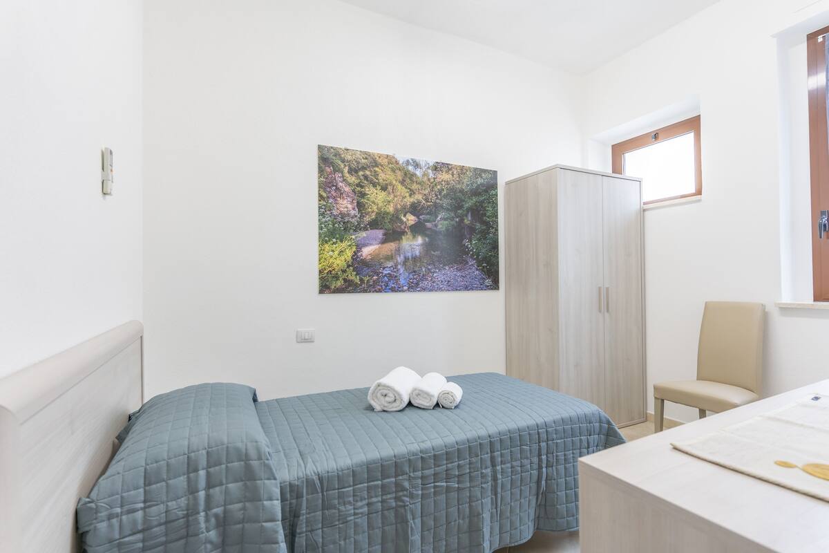 A bedroom with a bed, a desk, a chair, a wardrobe, and a painting of a landscape.