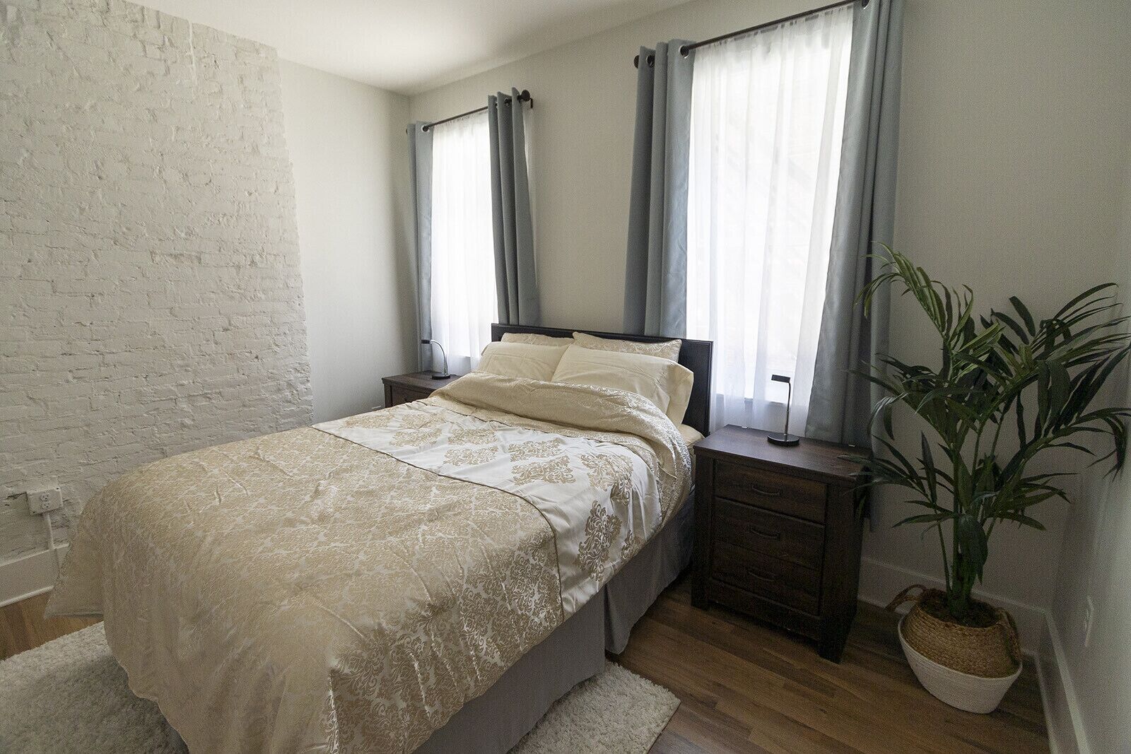 Un dormitorio con cama, mesitas de noche, una planta en maceta y una pared de ladrillo blanco.