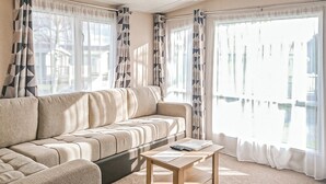 A living room with a sofa, a coffee table, and large windows with curtains. - York Standard Caravan (Thirsk)