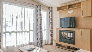 A living room with a television on a wooden cabinet, a fireplace, and a table with a remote control. - York Standard Caravan (Thirsk)
