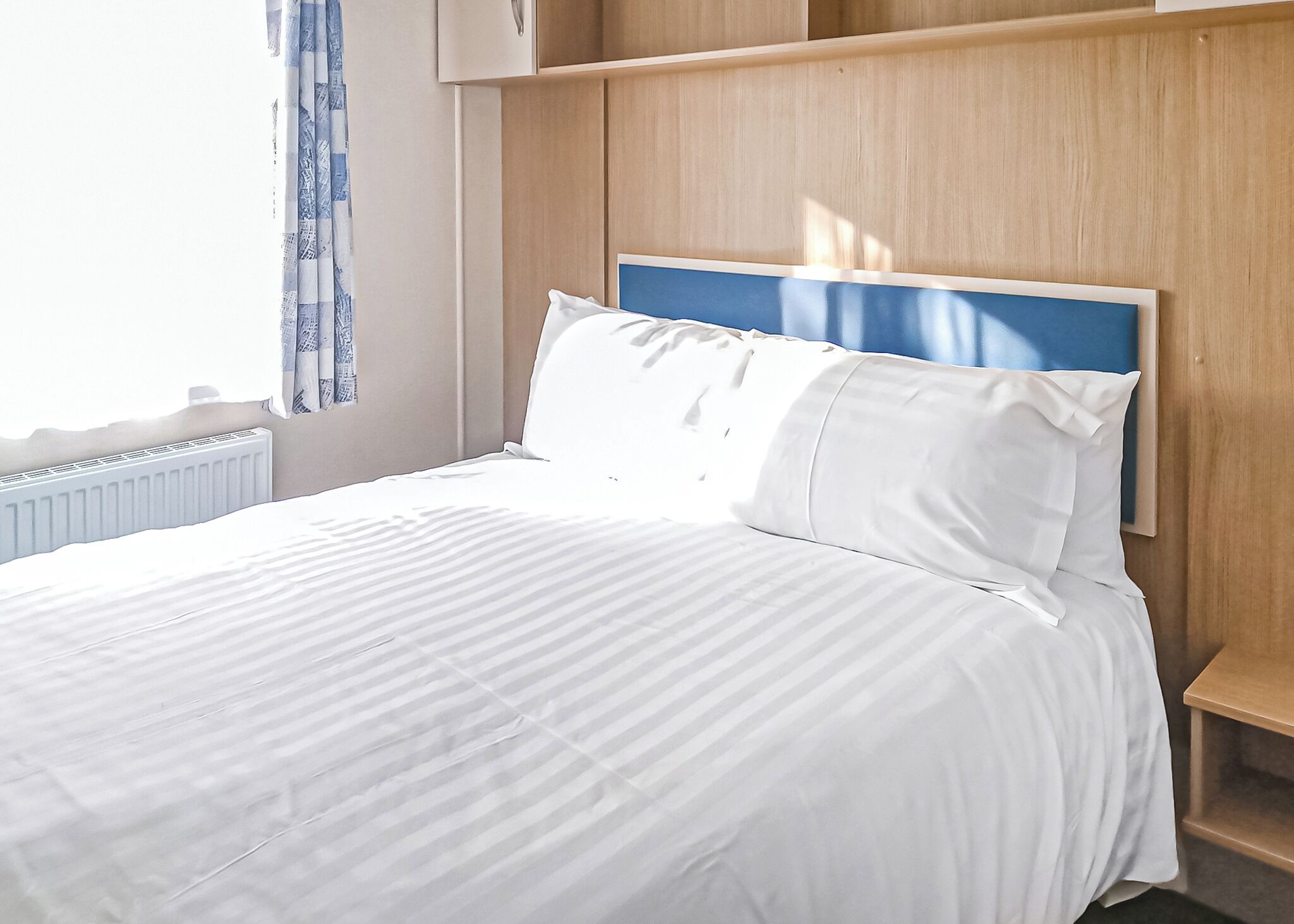 Een net opgemaakt bed met witte lakens, een houten hoofdbord en een raam met blauwe gordijnen.