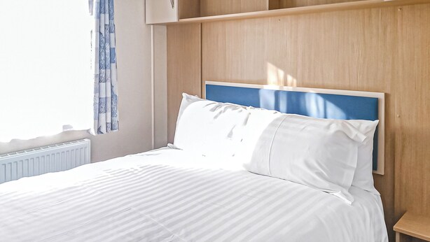 A neatly made bed with white bedding, a wooden headboard, and a window with blue curtains. - York Standard Caravan (Thirsk)