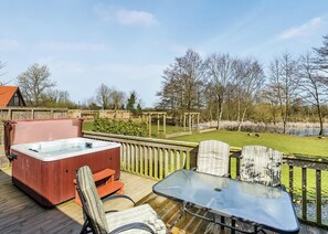 Une terrasse dotée d’un jacuzzi, de chaises et d’une table, offrant une vue sur un étang et des arbres.