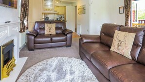 Ein Wohnzimmer mit Kamin, zwei Ledersofas, einem weißen Teppich und einer Küche im Hintergrund.