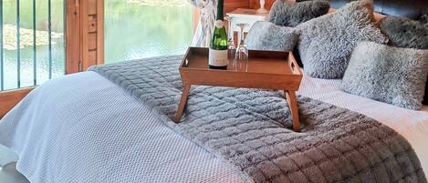 Quarto de cabana de madeira com cama, travesseiros, mesa de cabeceira com abajur e vista para um lago.