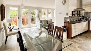 A modern kitchen with a glass dining table, wooden chairs, and a view of the garden through large sliding doors. - Country Lodge Four Platinum (Tintagel)