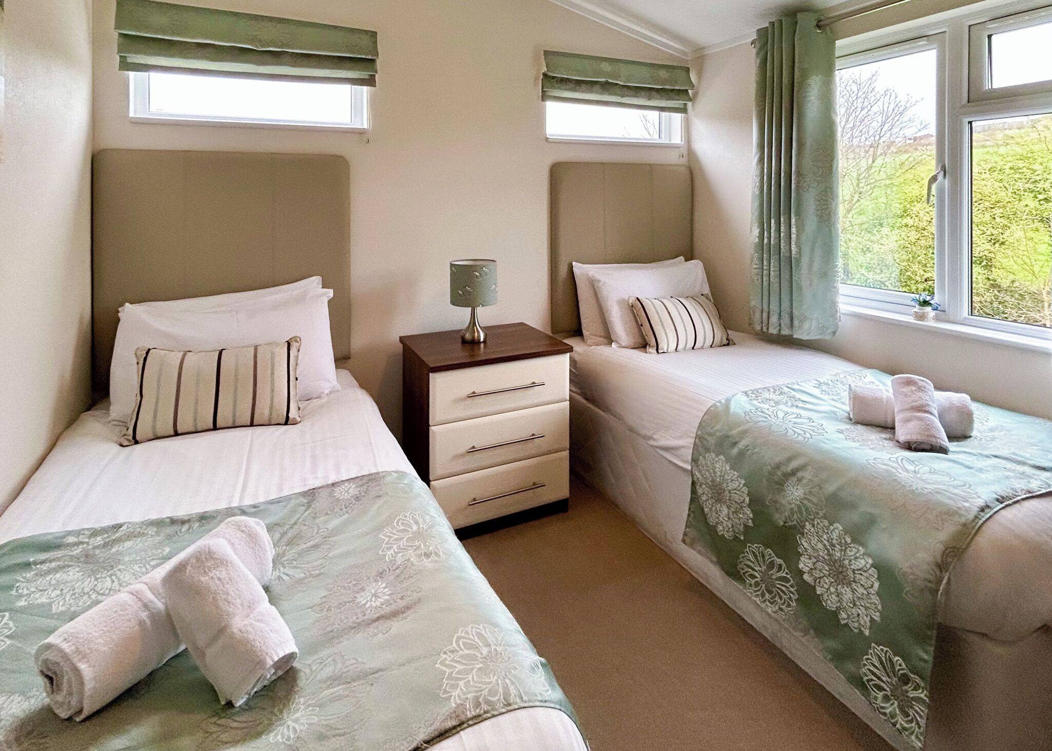 A hotel room with two single beds, a nightstand with a lamp, a dresser, and a window with curtains.