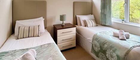 A hotel room with two single beds, a nightstand with a lamp, a dresser, and a window with curtains.