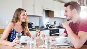 Un couple est assis à une table dans une cuisine moderne, avec des meubles blancs, une cuisinière et une bouilloire.