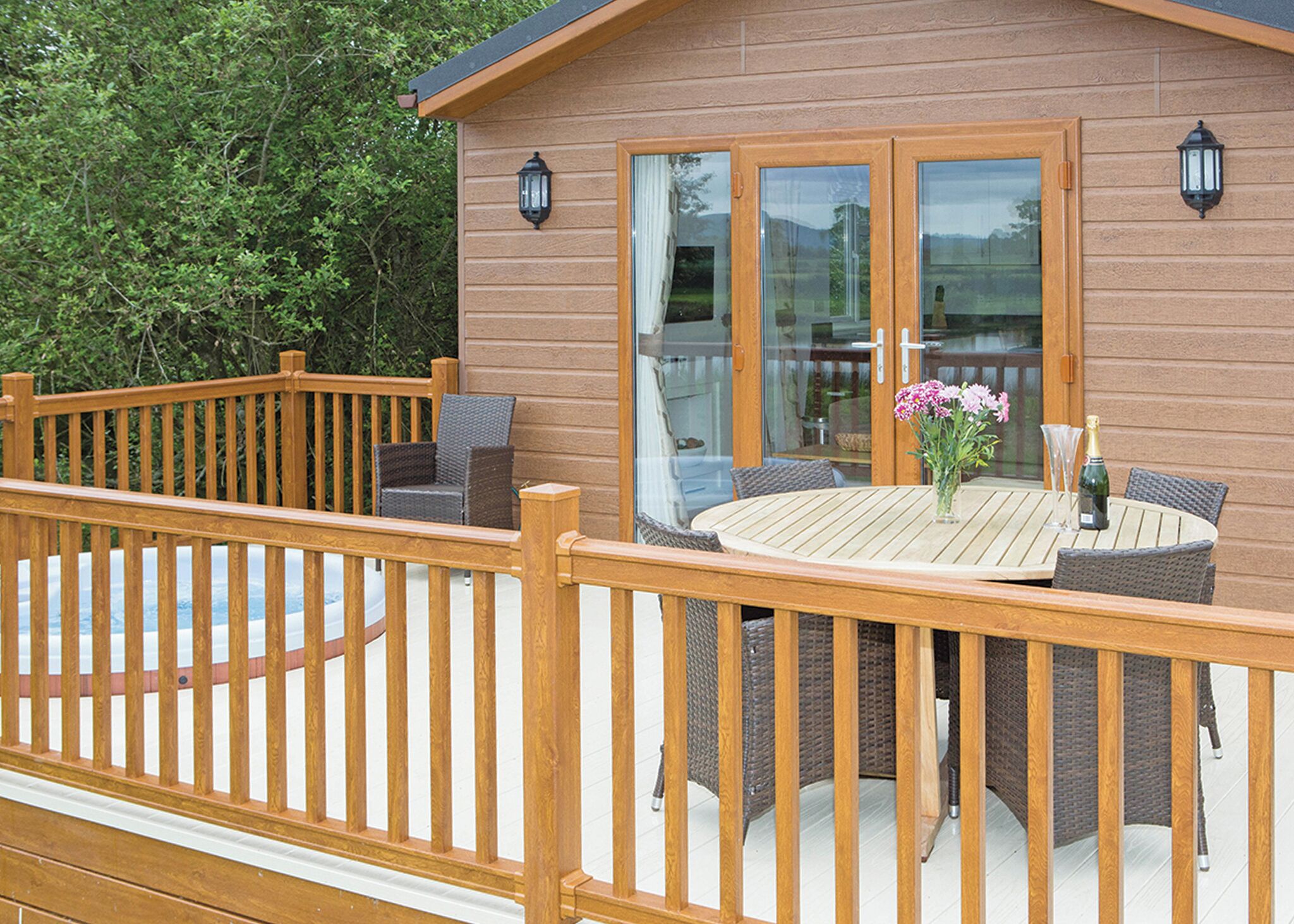 A wooden deck with a round table and wicker chairs, a glass door, and two wall-mounted lanterns.