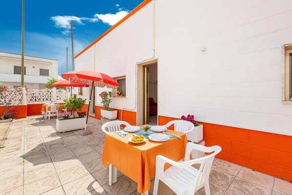 An outdoor patio with a table set for dining, surrounded by white chairs and a red umbrella.
