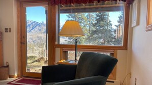 Une pièce avec vue sur les montagnes, visible à travers une grande fenêtre, une lampe sur une table d’appoint et une chaise au premier plan.