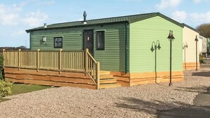 A green mobile home with a wooden deck, a door, and two exterior wall-mounted lights.