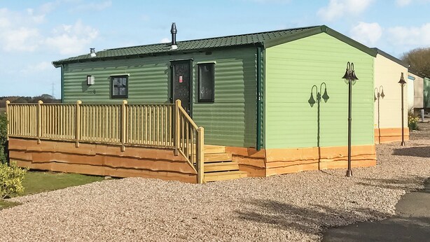 A green mobile home with a wooden deck, a door, and two exterior wall-mounted lights. - Caravan with one double and one twin. (Cargill)