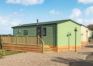 A green mobile home with a wooden deck, a door, and two exterior wall-mounted lights.