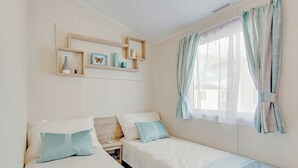 A bedroom with two beds, a window with sheer curtains, and a shelf with decorative items. - Caravan with one double and one twin. (Cargill)
