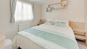 A bedroom with a bed, a nightstand, a shelf with decorative items, and a window with curtains. - Caravan with one double and one twin. (Cargill)