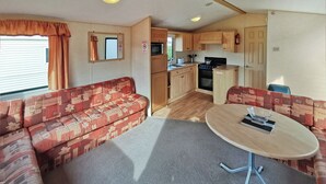 A compact RV interior with a sofa, a dining table, a kitchen area, and a window with curtains. - 2 bedroom accommodation in Clarach Bay, Nr Aberystwyth (Clarach Bay, Nr Aberystwyth)