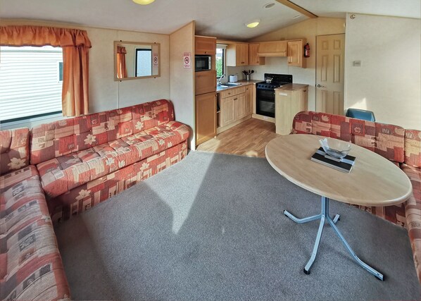 A compact RV interior with a sofa, a dining table, a kitchen area, and a window with curtains.
