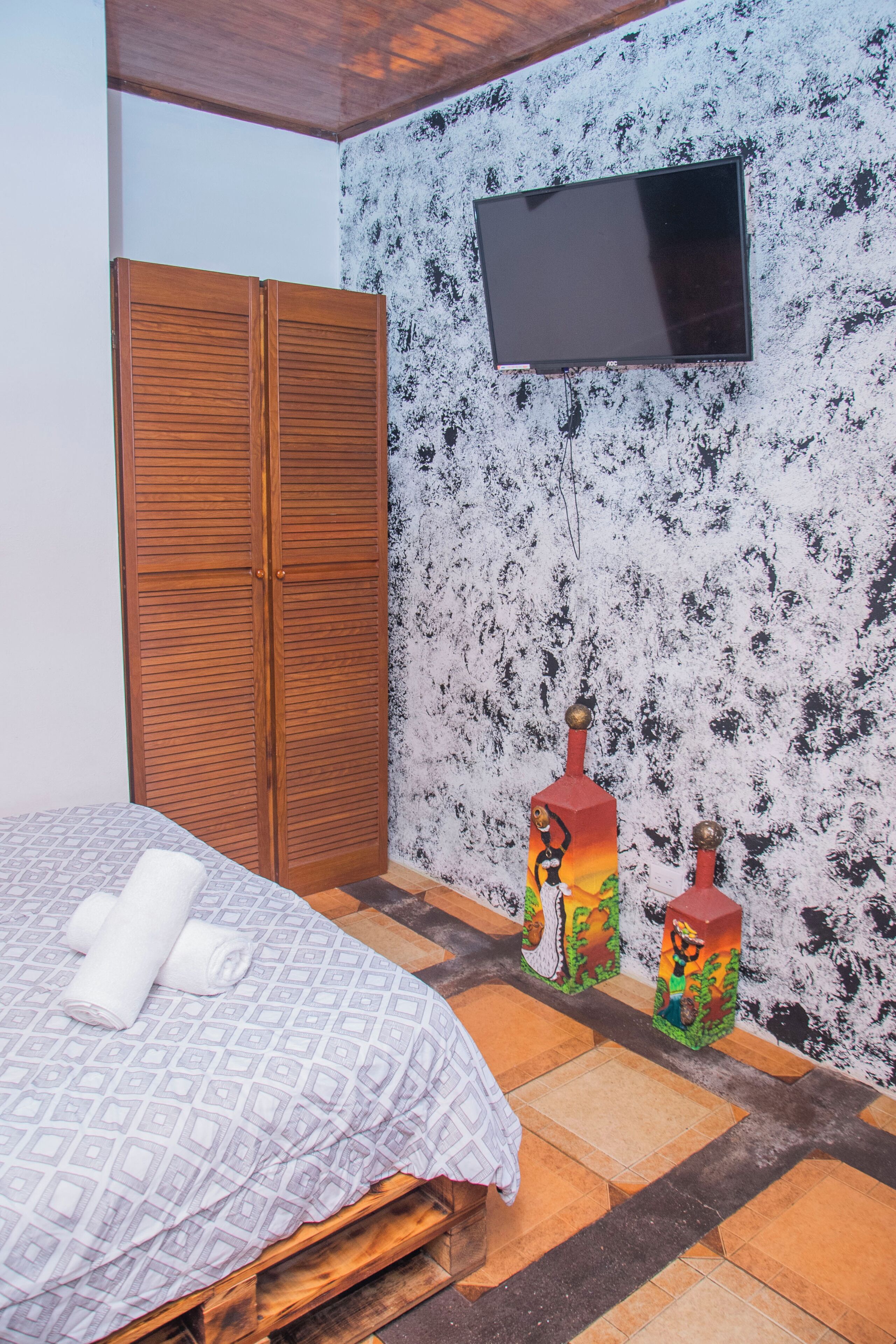 A bedroom with a bed, wooden wardrobe, a flat-screen TV, and decorative bottles on the wall.