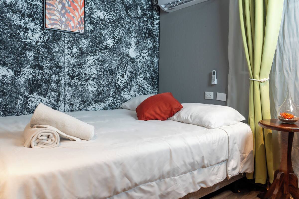 A neatly made bed with white and red pillows, a rolled white towel, and a small wooden side table with a glass vase and a bowl of fruit.