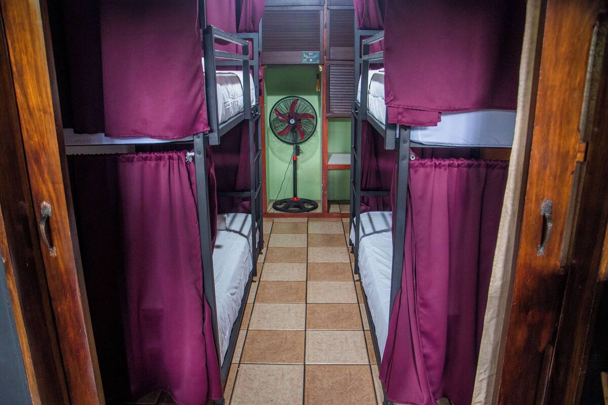 A narrow corridor with bunk beds on either side, a fan in the center, and a green wall.
