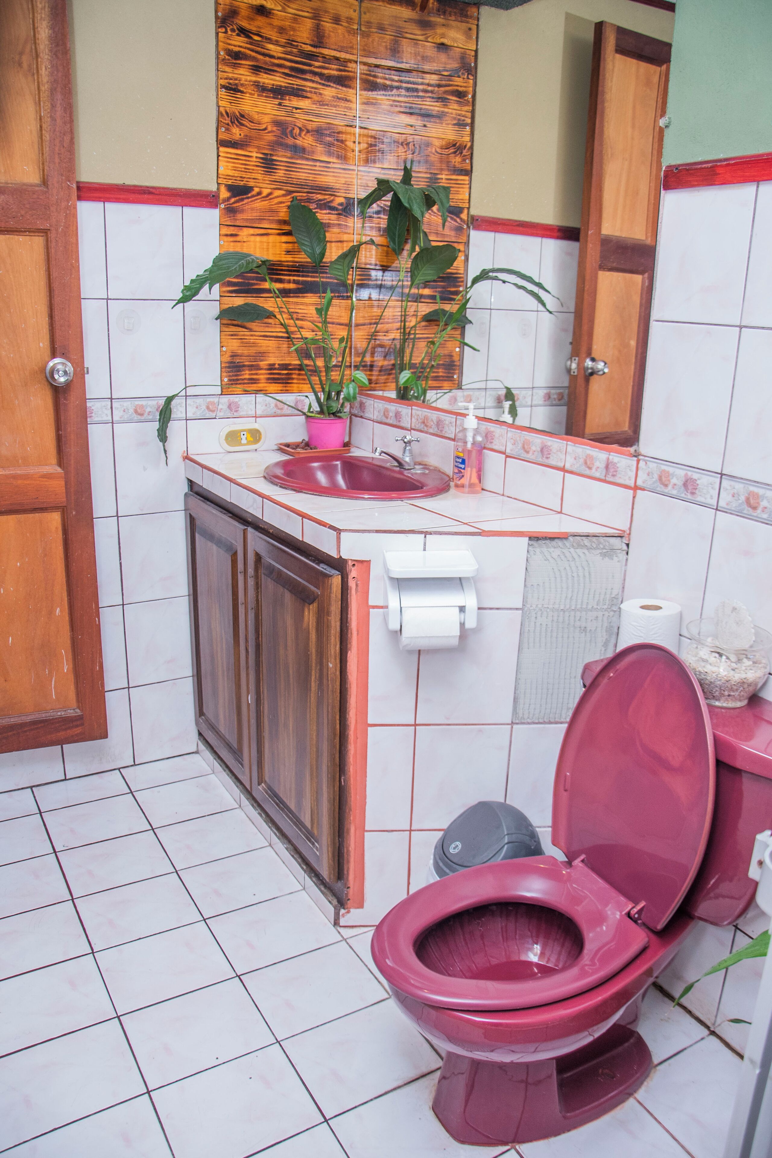 Een badkamer met een rood toilet, een houten kastje, een wastafel met een roze zeepdispenser en een spiegel boven de wastafel.