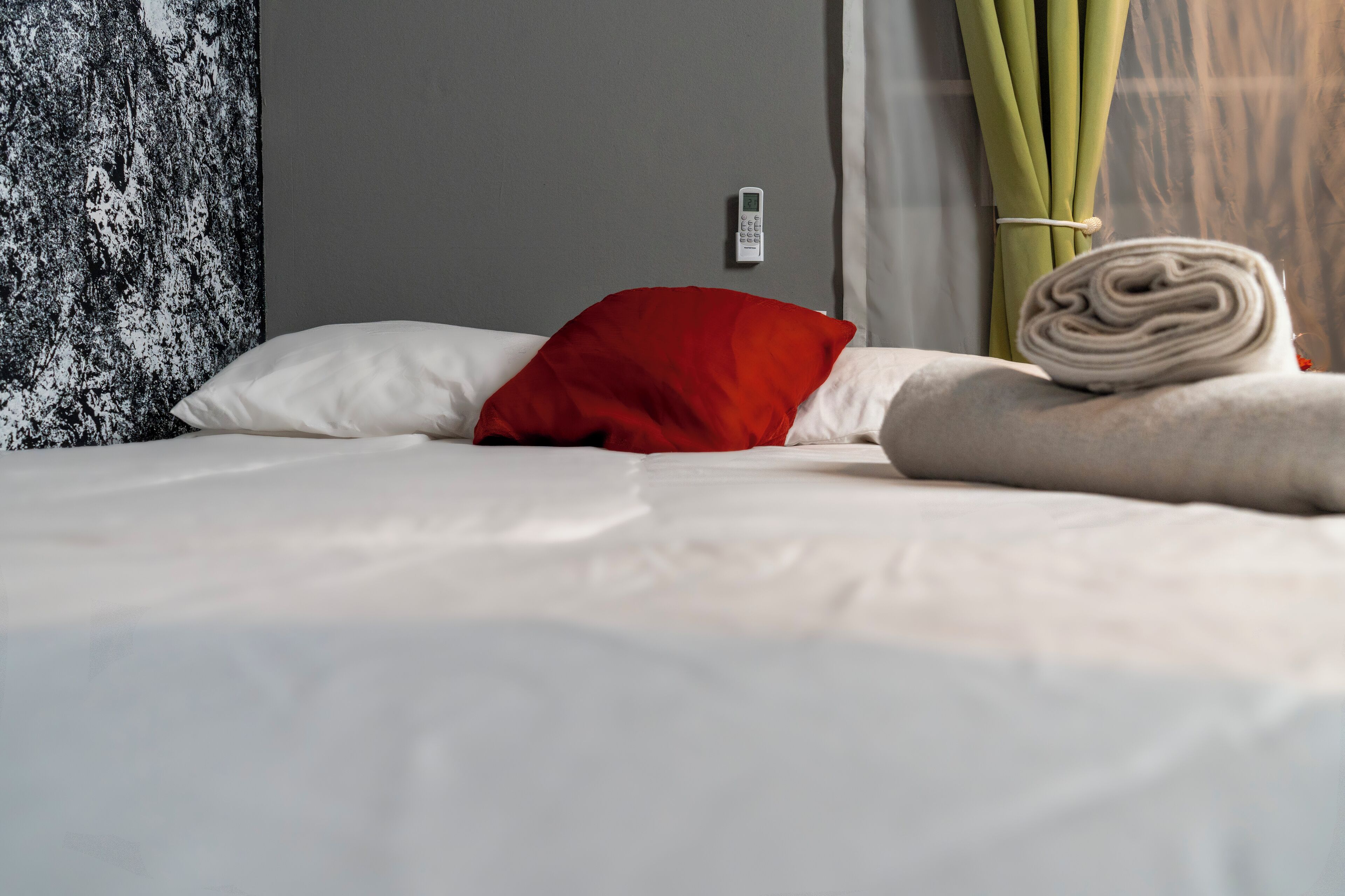 A neatly made bed with white and red pillows, a rolled-up towel, and a wall-mounted thermostat.