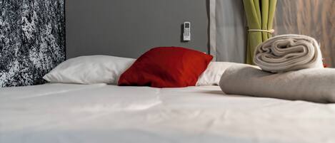 A neatly made bed with white and red pillows, a rolled-up towel, and a wall-mounted thermostat.