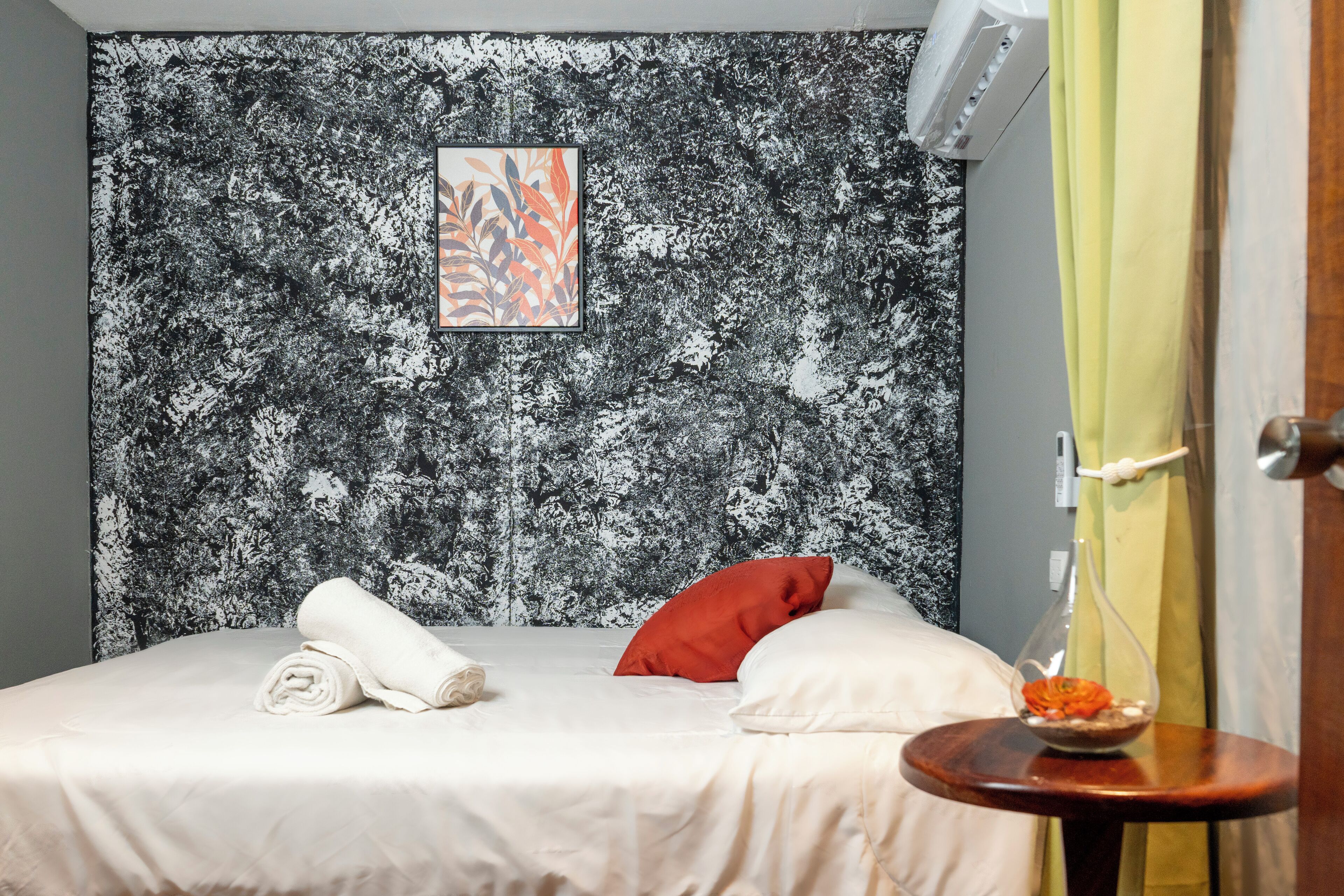 A hotel room with a bed, a towel, a pillow, a small table with a vase, and a wall with a textured pattern.